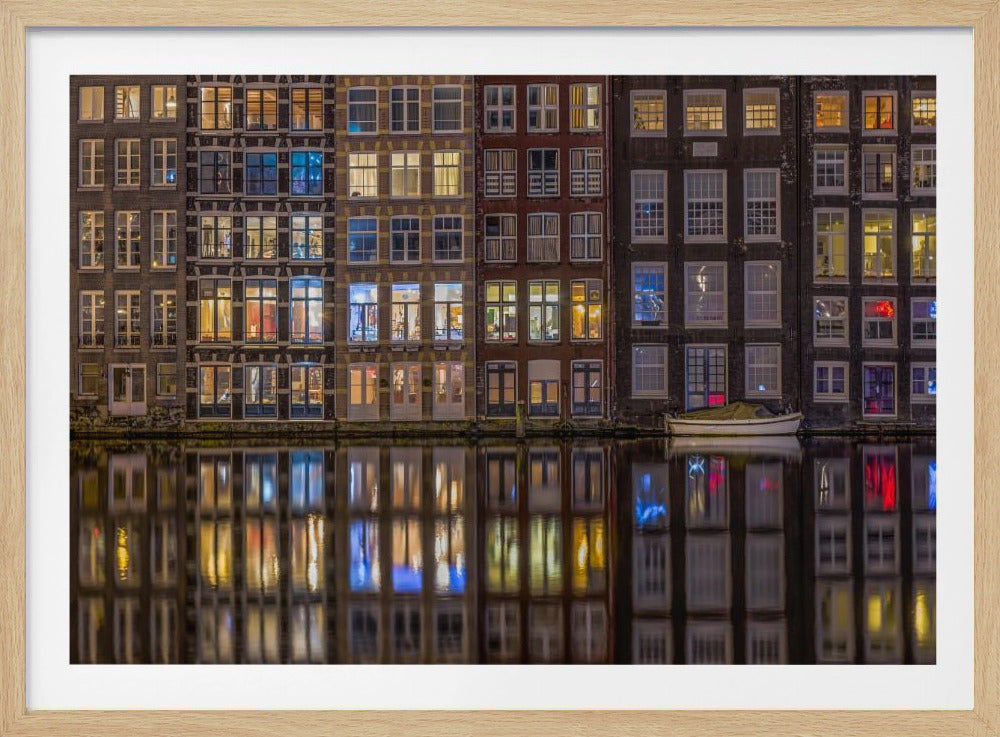 A framed nighttime photograph of a row of traditional Amsterdam canal houses with many illuminated windows, their colorful lights reflecting perfectly in the calm canal water below. A small white boat is moored at the water's edge. Decor