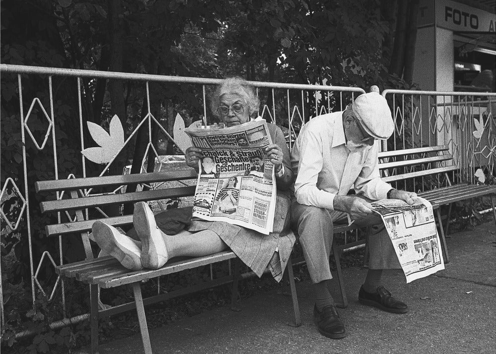 A framed black and white photograph of an elderly couple sitting on a park bench, both deeply absorbed in reading their newspapers. The woman on the left has her feet propped up on the bench, while the man on the right sits next to her, looking down at his paper. Poster