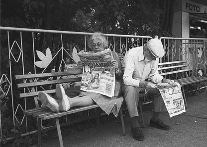 A framed black and white photograph of an elderly couple sitting on a park bench, both deeply absorbed in reading their newspapers. The woman on the left has her feet propped up on the bench, while the man on the right sits next to her, looking down at his paper. Poster