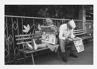 A framed black and white photograph of an elderly couple sitting on a park bench, both deeply absorbed in reading their newspapers. The woman on the left has her feet propped up on the bench, while the man on the right sits next to her, looking down at his paper. Poster