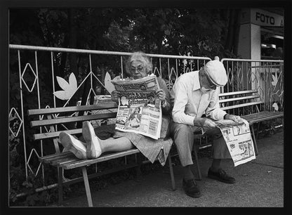 A framed black and white photograph of an elderly couple sitting on a park bench, both deeply absorbed in reading their newspapers. The woman on the left has her feet propped up on the bench, while the man on the right sits next to her, looking down at his paper. Poster
