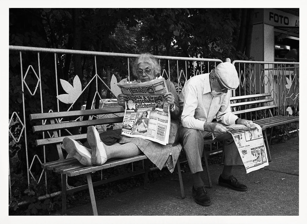A framed black and white photograph of an elderly couple sitting on a park bench, both deeply absorbed in reading their newspapers. The woman on the left has her feet propped up on the bench, while the man on the right sits next to her, looking down at his paper. Poster