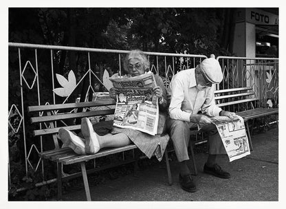 A framed black and white photograph of an elderly couple sitting on a park bench, both deeply absorbed in reading their newspapers. The woman on the left has her feet propped up on the bench, while the man on the right sits next to her, looking down at his paper. Poster