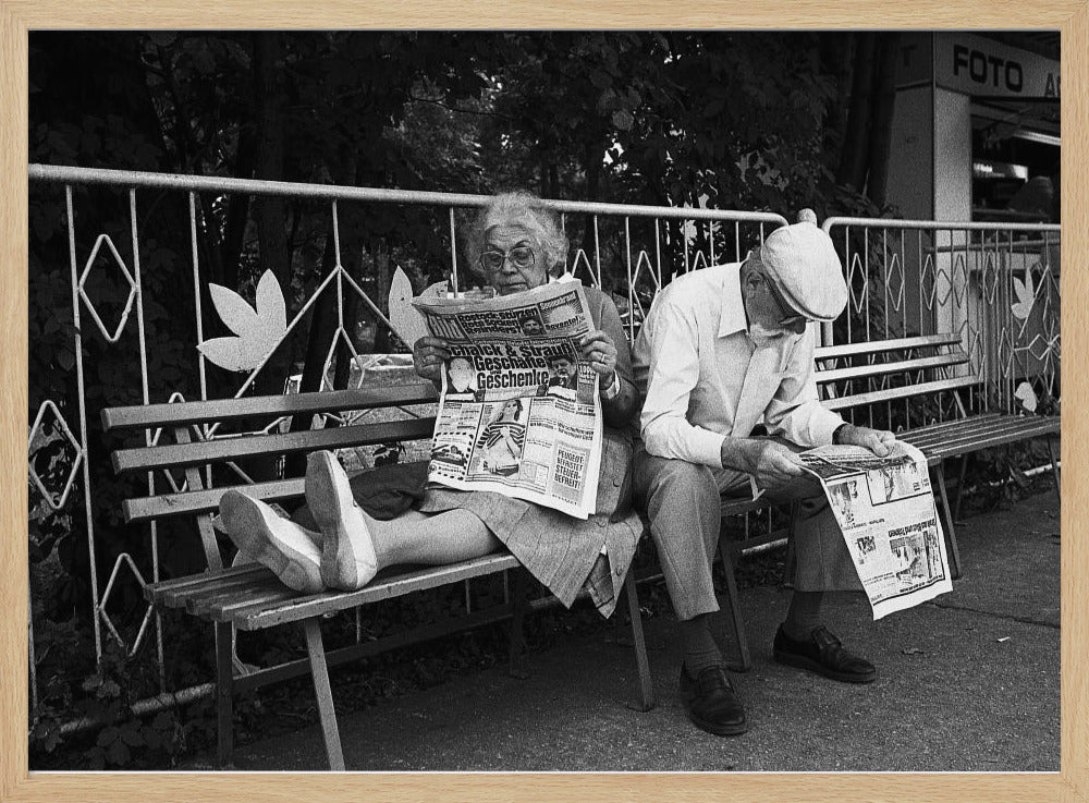 A framed black and white photograph of an elderly couple sitting on a park bench, both deeply absorbed in reading their newspapers. The woman on the left has her feet propped up on the bench, while the man on the right sits next to her, looking down at his paper. Poster