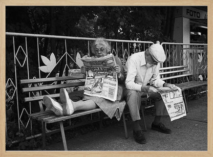 A framed black and white photograph of an elderly couple sitting on a park bench, both deeply absorbed in reading their newspapers. The woman on the left has her feet propped up on the bench, while the man on the right sits next to her, looking down at his paper. Poster