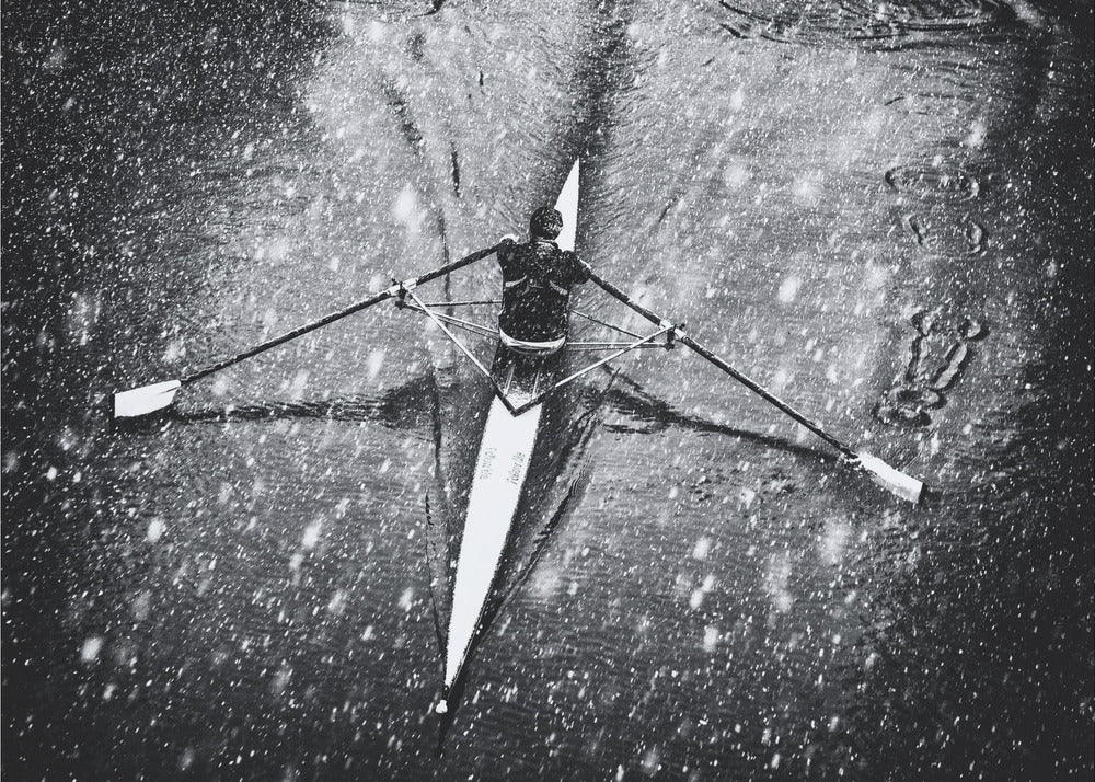 A dramatic black and white overhead photograph of a single rower in a narrow scull, captured mid-stroke as they cut through dark water during a heavy snowfall, creating a high-contrast and atmospheric scene. Decor