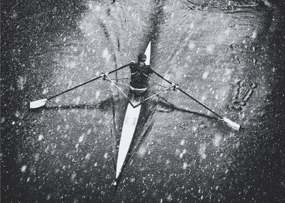 A dramatic black and white overhead photograph of a single rower in a narrow scull, captured mid-stroke as they cut through dark water during a heavy snowfall, creating a high-contrast and atmospheric scene. Decor