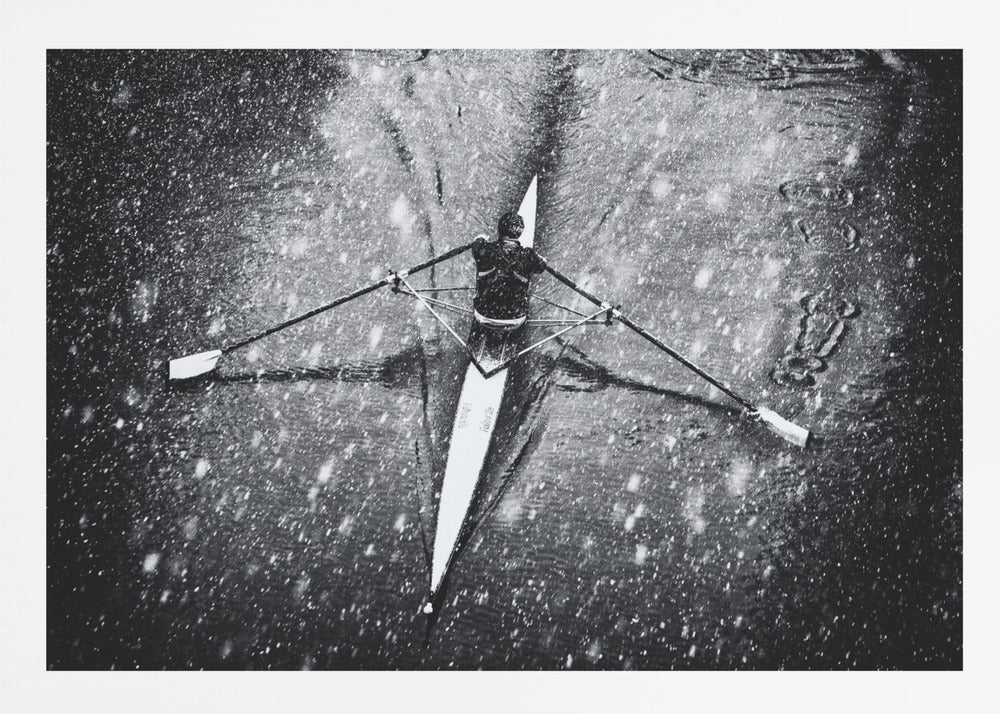 A dramatic black and white overhead photograph of a single rower in a narrow scull, captured mid-stroke as they cut through dark water during a heavy snowfall, creating a high-contrast and atmospheric scene. Decor