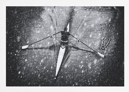 A dramatic black and white overhead photograph of a single rower in a narrow scull, captured mid-stroke as they cut through dark water during a heavy snowfall, creating a high-contrast and atmospheric scene. Decor