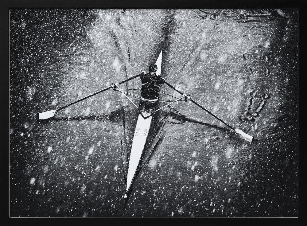 A dramatic black and white overhead photograph of a single rower in a narrow scull, captured mid-stroke as they cut through dark water during a heavy snowfall, creating a high-contrast and atmospheric scene. Decor