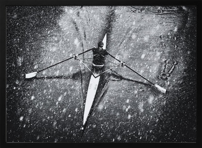 A dramatic black and white overhead photograph of a single rower in a narrow scull, captured mid-stroke as they cut through dark water during a heavy snowfall, creating a high-contrast and atmospheric scene. Decor