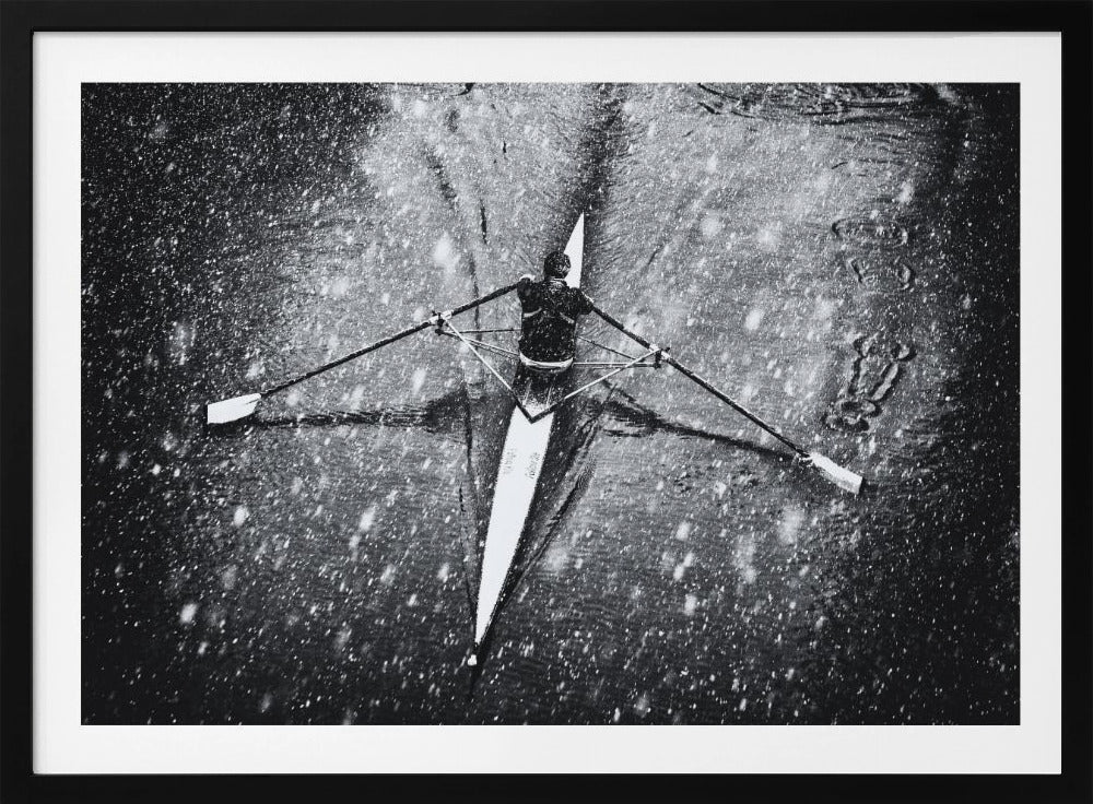 A dramatic black and white overhead photograph of a single rower in a narrow scull, captured mid-stroke as they cut through dark water during a heavy snowfall, creating a high-contrast and atmospheric scene. Decor