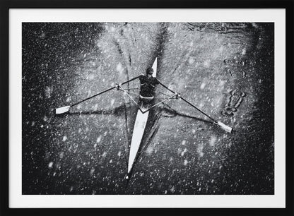 A dramatic black and white overhead photograph of a single rower in a narrow scull, captured mid-stroke as they cut through dark water during a heavy snowfall, creating a high-contrast and atmospheric scene. Decor