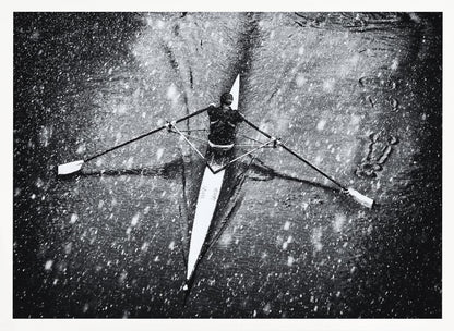 A dramatic black and white overhead photograph of a single rower in a narrow scull, captured mid-stroke as they cut through dark water during a heavy snowfall, creating a high-contrast and atmospheric scene. Decor