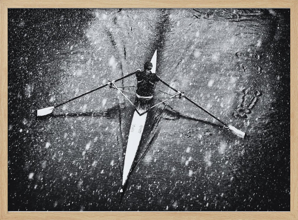 A dramatic black and white overhead photograph of a single rower in a narrow scull, captured mid-stroke as they cut through dark water during a heavy snowfall, creating a high-contrast and atmospheric scene. Decor