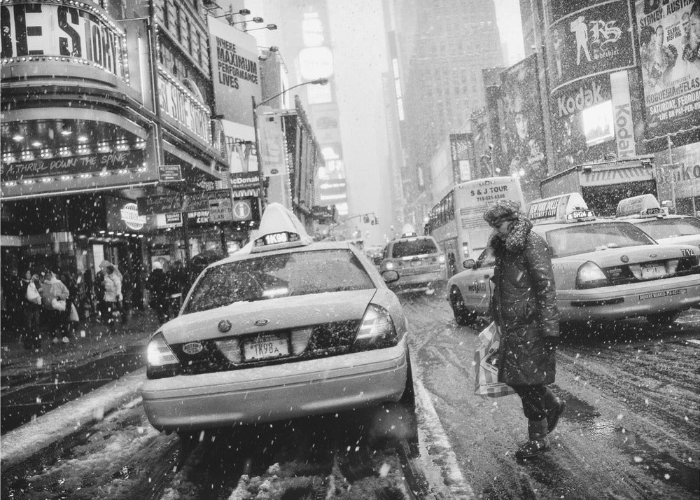 A black and white photograph of a bustling New York City street during a snowstorm. A person in a heavy winter coat crosses the street in front of a line of yellow cabs, with the bright lights of Times Square billboards glowing softly in the background through the falling snow. Print