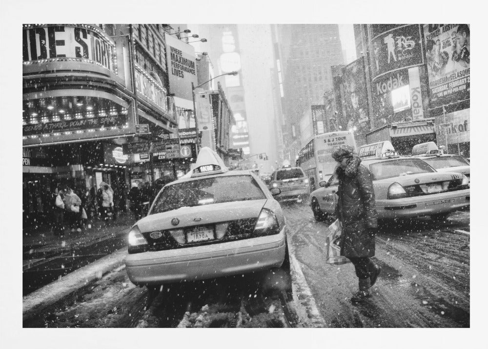 A black and white photograph of a bustling New York City street during a snowstorm. A person in a heavy winter coat crosses the street in front of a line of yellow cabs, with the bright lights of Times Square billboards glowing softly in the background through the falling snow. Print