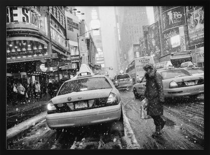 A black and white photograph of a bustling New York City street during a snowstorm. A person in a heavy winter coat crosses the street in front of a line of yellow cabs, with the bright lights of Times Square billboards glowing softly in the background through the falling snow. Print