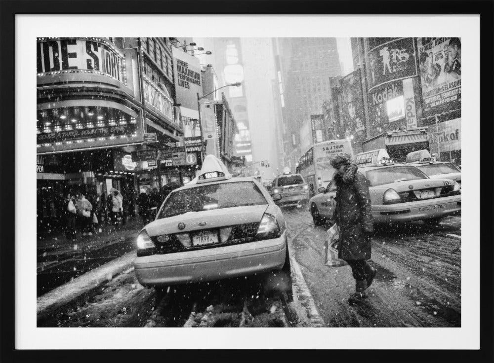 A black and white photograph of a bustling New York City street during a snowstorm. A person in a heavy winter coat crosses the street in front of a line of yellow cabs, with the bright lights of Times Square billboards glowing softly in the background through the falling snow. Print