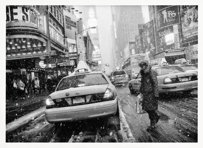 A black and white photograph of a bustling New York City street during a snowstorm. A person in a heavy winter coat crosses the street in front of a line of yellow cabs, with the bright lights of Times Square billboards glowing softly in the background through the falling snow. Print