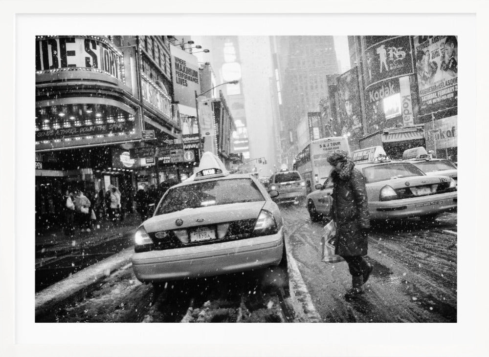 A black and white photograph of a bustling New York City street during a snowstorm. A person in a heavy winter coat crosses the street in front of a line of yellow cabs, with the bright lights of Times Square billboards glowing softly in the background through the falling snow. Print