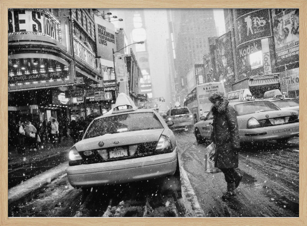 A black and white photograph of a bustling New York City street during a snowstorm. A person in a heavy winter coat crosses the street in front of a line of yellow cabs, with the bright lights of Times Square billboards glowing softly in the background through the falling snow. Print
