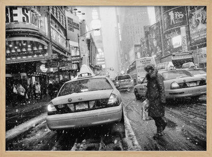 A black and white photograph of a bustling New York City street during a snowstorm. A person in a heavy winter coat crosses the street in front of a line of yellow cabs, with the bright lights of Times Square billboards glowing softly in the background through the falling snow. Print