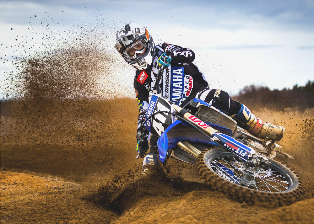 A motocross rider in full gear leans sharply into a turn on a dirt track, kicking up a large spray of mud and dirt from their blue and white Yamaha motorcycle. Artwork