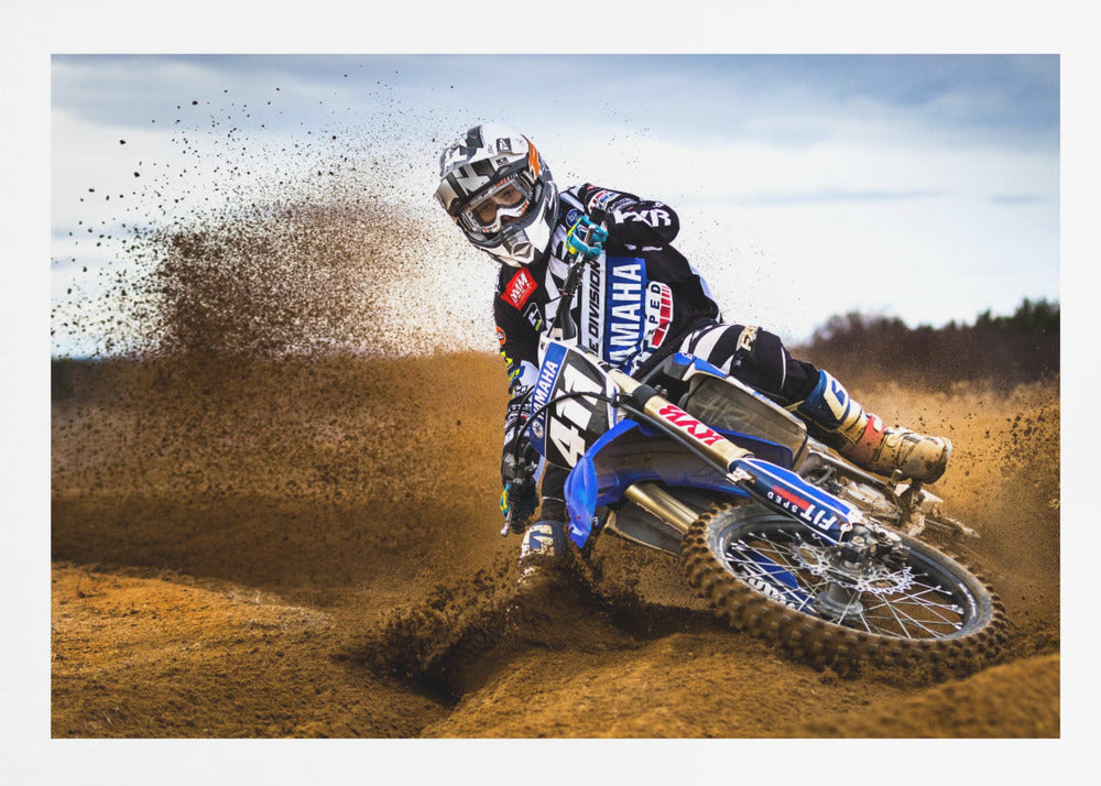 A motocross rider in full gear leans sharply into a turn on a dirt track, kicking up a large spray of mud and dirt from their blue and white Yamaha motorcycle. Artwork