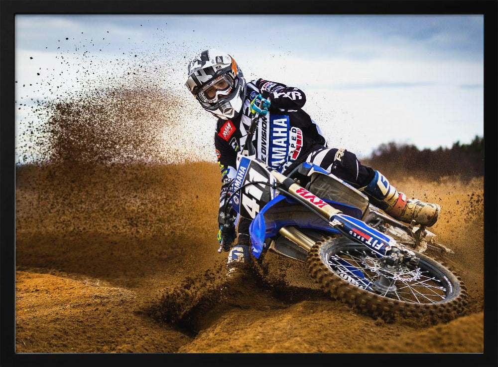A motocross rider in full gear leans sharply into a turn on a dirt track, kicking up a large spray of mud and dirt from their blue and white Yamaha motorcycle. Artwork