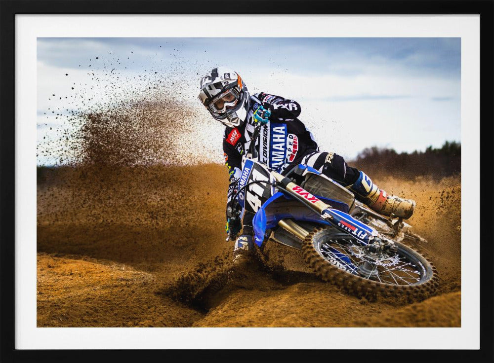 A motocross rider in full gear leans sharply into a turn on a dirt track, kicking up a large spray of mud and dirt from their blue and white Yamaha motorcycle. Artwork