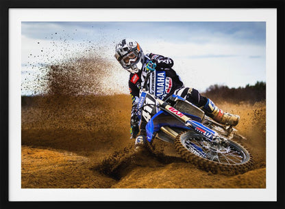 A motocross rider in full gear leans sharply into a turn on a dirt track, kicking up a large spray of mud and dirt from their blue and white Yamaha motorcycle. Artwork
