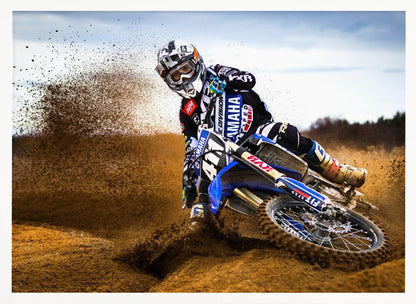 A motocross rider in full gear leans sharply into a turn on a dirt track, kicking up a large spray of mud and dirt from their blue and white Yamaha motorcycle. Artwork