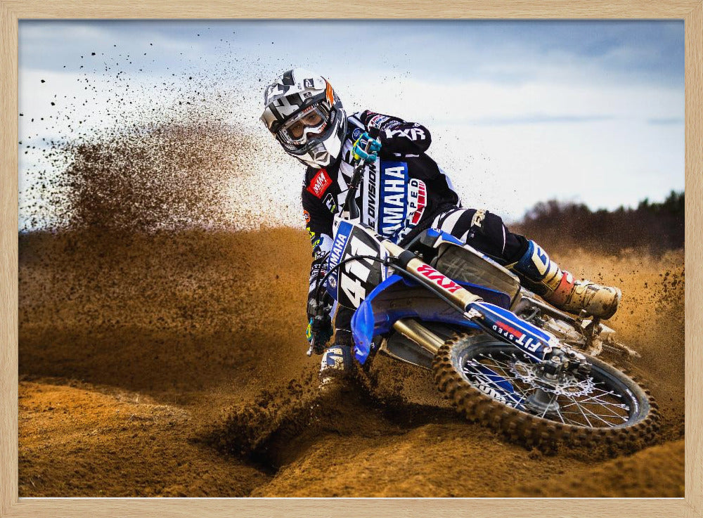 A motocross rider in full gear leans sharply into a turn on a dirt track, kicking up a large spray of mud and dirt from their blue and white Yamaha motorcycle. Artwork