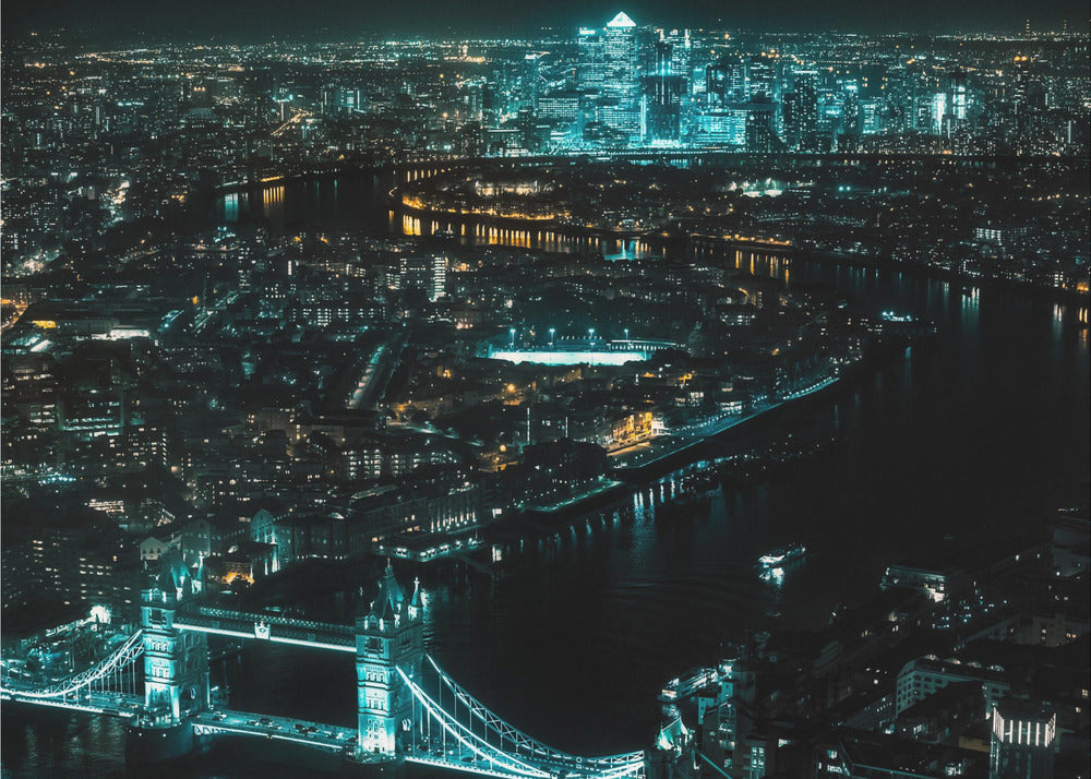 A framed aerial photograph of London at night, featuring the iconic Tower Bridge illuminated in bright teal in the foreground and the glowing skyscrapers of Canary Wharf across the River Thames in the background. Artwork