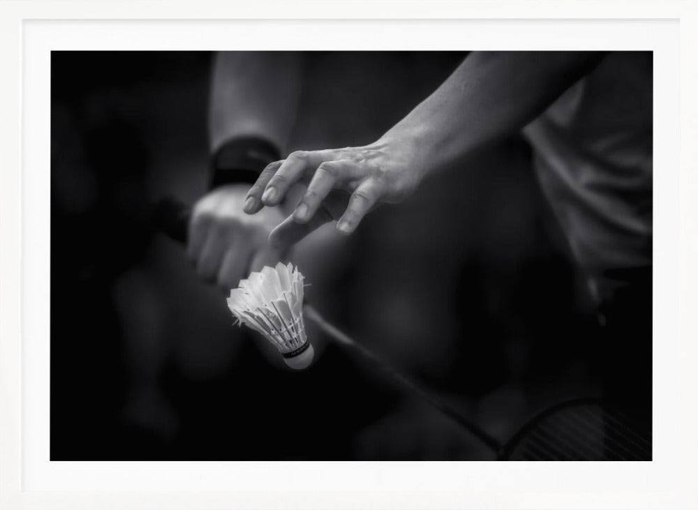 A dramatic black and white close-up photograph of a badminton player preparing to serve. The focus is on the player's hands, with one hand delicately positioned above a shuttlecock which is balanced on the strings of a racquet. Artwork