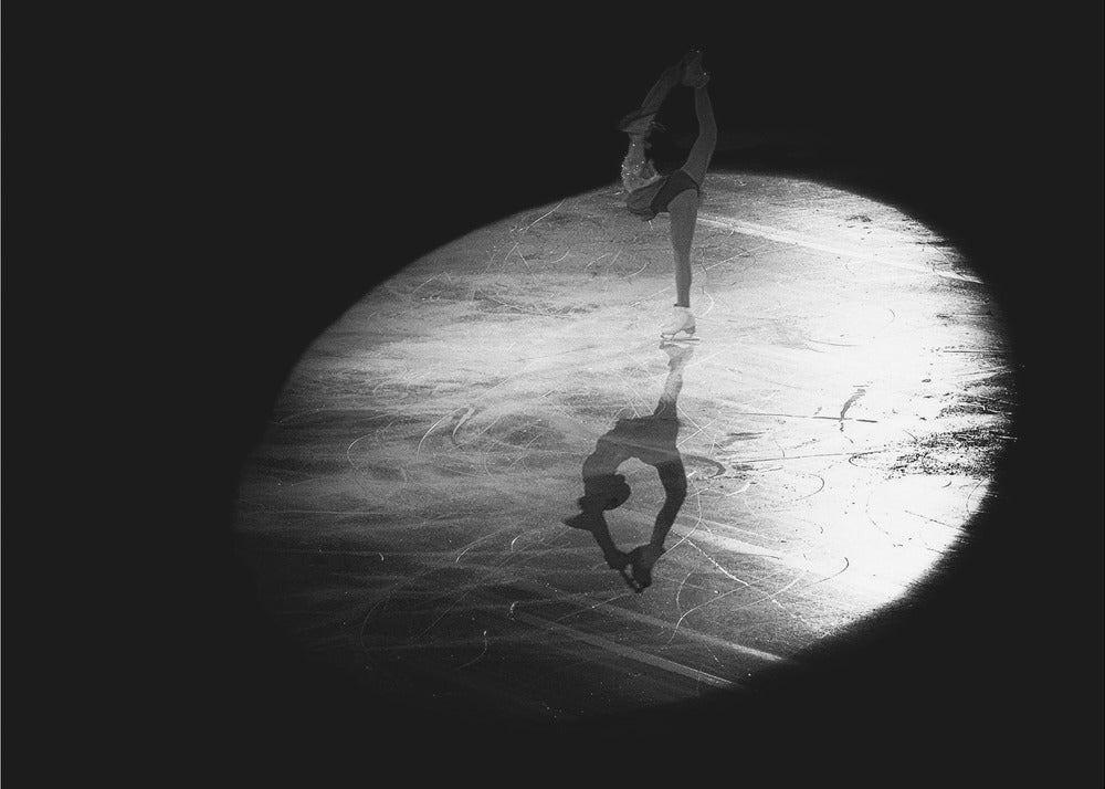 A dramatic black and white framed photograph of a female figure skater performing on ice. She is illuminated by a single spotlight, holding a graceful standing split pose, with her reflection visible on the ice below. Poster