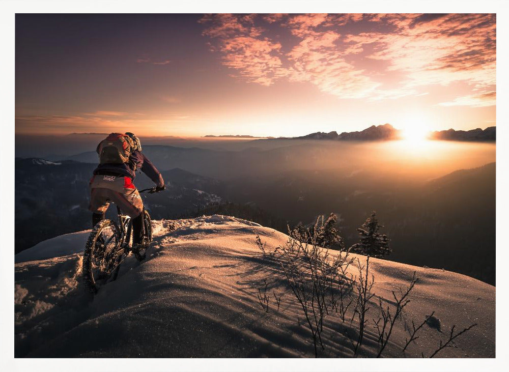 A mountain biker seen from behind pauses on a snowy mountain summit, overlooking a vast, misty mountain range as the sun sets, casting a warm orange and pink glow across the sky and snow. Artwork