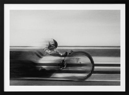 A black and white photograph captured with motion blur, showing a cyclist in an aerodynamic position racing at high speed. The background is blurred into horizontal lines, emphasizing the velocity. The image is enclosed in a silver frame. Wall Art