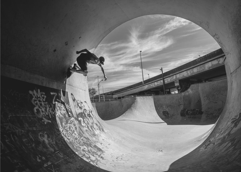 A dynamic black and white action shot of a skateboarder mid-trick on the edge of a concrete ramp in a graffiti-covered skatepark, framed by the curve of a large pipe. Print