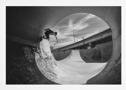 A dynamic black and white action shot of a skateboarder mid-trick on the edge of a concrete ramp in a graffiti-covered skatepark, framed by the curve of a large pipe. Print