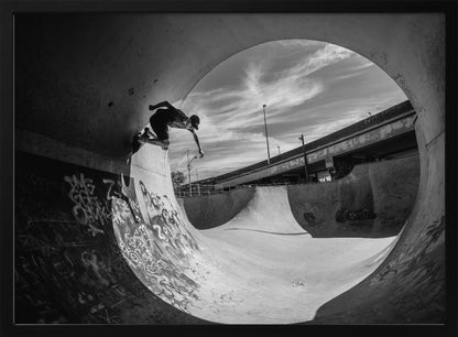 A dynamic black and white action shot of a skateboarder mid-trick on the edge of a concrete ramp in a graffiti-covered skatepark, framed by the curve of a large pipe. Print