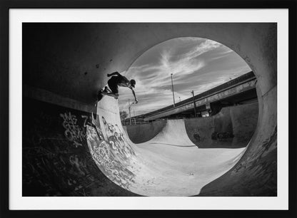 A dynamic black and white action shot of a skateboarder mid-trick on the edge of a concrete ramp in a graffiti-covered skatepark, framed by the curve of a large pipe. Print