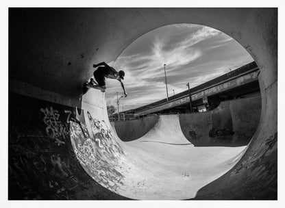 A dynamic black and white action shot of a skateboarder mid-trick on the edge of a concrete ramp in a graffiti-covered skatepark, framed by the curve of a large pipe. Print