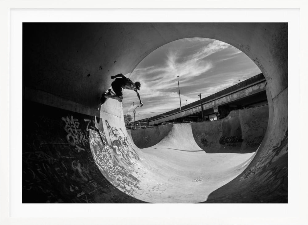 A dynamic black and white action shot of a skateboarder mid-trick on the edge of a concrete ramp in a graffiti-covered skatepark, framed by the curve of a large pipe. Print