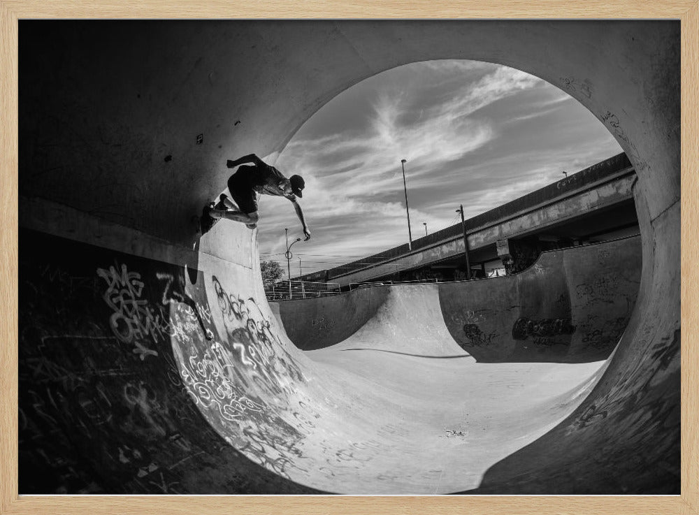 A dynamic black and white action shot of a skateboarder mid-trick on the edge of a concrete ramp in a graffiti-covered skatepark, framed by the curve of a large pipe. Print