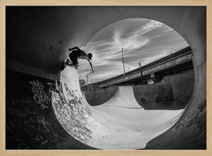A dynamic black and white action shot of a skateboarder mid-trick on the edge of a concrete ramp in a graffiti-covered skatepark, framed by the curve of a large pipe. Print