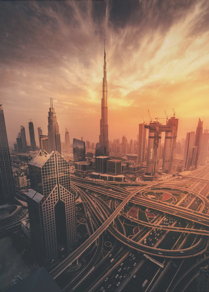 A dramatic high-angle photograph of the Dubai skyline at sunset. The Burj Khalifa stands tall in the center, bathed in the warm light of the setting sun, while a complex network of highways and interchanges weaves between modern skyscrapers in the foreground under a cloudy, orange-tinted sky. Poster