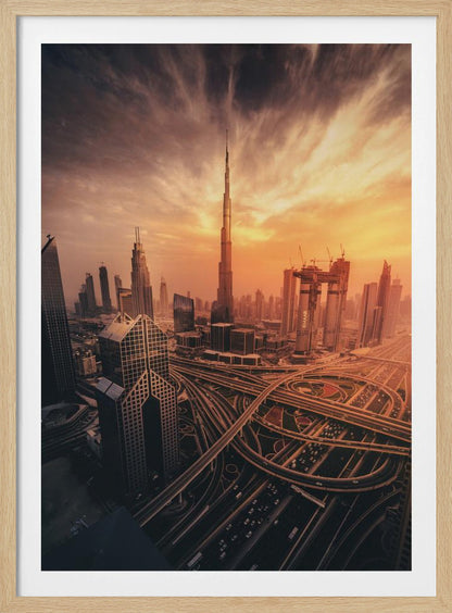 A dramatic high-angle photograph of the Dubai skyline at sunset. The Burj Khalifa stands tall in the center, bathed in the warm light of the setting sun, while a complex network of highways and interchanges weaves between modern skyscrapers in the foreground under a cloudy, orange-tinted sky. Poster