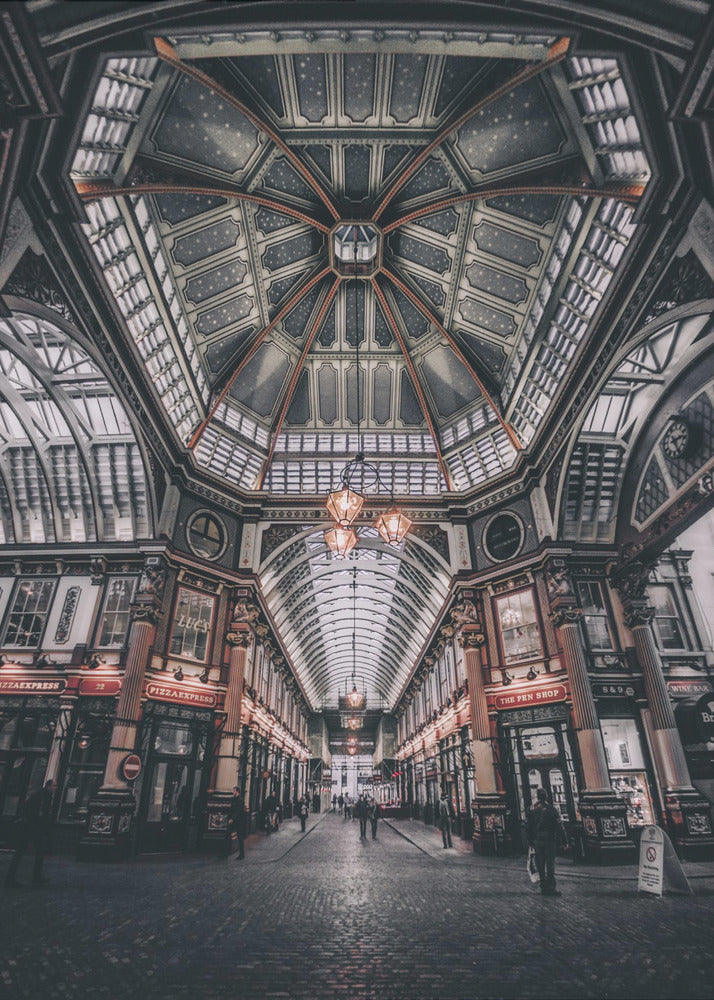 A wide-angle, symmetrical photograph looking down the center of an ornate Victorian covered market, Leadenhall Market. The focus is on the elaborate, arched glass and ironwork ceiling which is decorated in dark blue with gold stars. Below, a cobblestone path is lined with traditional shops with glowing lights, and a few people can be seen walking in the distance. The image has a moody, high-contrast aesthetic. Wall Art